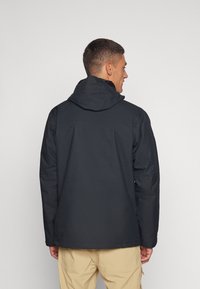 Veste noire imperméable avec capuche, présentant une texture lisse et une coupe légèrement ample, montrée de dos sur fond uni.