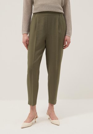 Femme portant un pantalon fuselé vert olive, un pull beige texturé et des escarpins slingback crème à bout pointu, debout devant un fond uni.