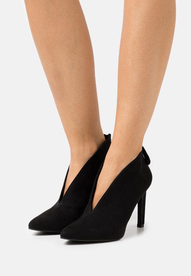 Jambes portant des bottines en daim noir à talons hauts, avec une coupe en V profonde à l'avant et des bouts pointus sur un fond blanc.