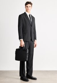 Czarny garnitur szyty na miarę z wełny, w połączeniu z białą koszulą i czarnym krawatem. Trzyma czarną skórzaną torbę z podwójnymi uchwytami, stojąc w czarnych butach.
