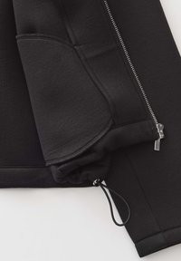 Zwarte sweatshirt met rits en een gestructureerde afwerking, voorzien van een kangoeroe-zoom en trekkoord bij de manchetten. Zilveren hardwareaccenten zichtbaar.