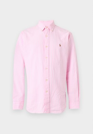 Mand iført hvid kasket, lyserød striped skjorte med knapper, lyseblå jeans med brunt bælte og sorte loafers, står med hænderne i lommerne.