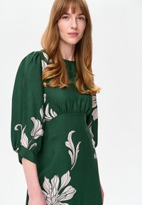 Zielona sukienka z lekkiego materiału, zdobiona dużymi białymi kwiatowymi wzorami. Rękawy balonowe z marszczeniami oraz dopasowana talia.