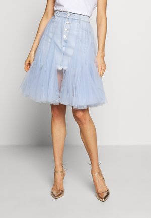 Femme portant une jupe en jean bleu clair boutonnée avec une superposition en tulle transparent et des talons métalliques à bride cheville à bout pointu, debout sur un fond uni.