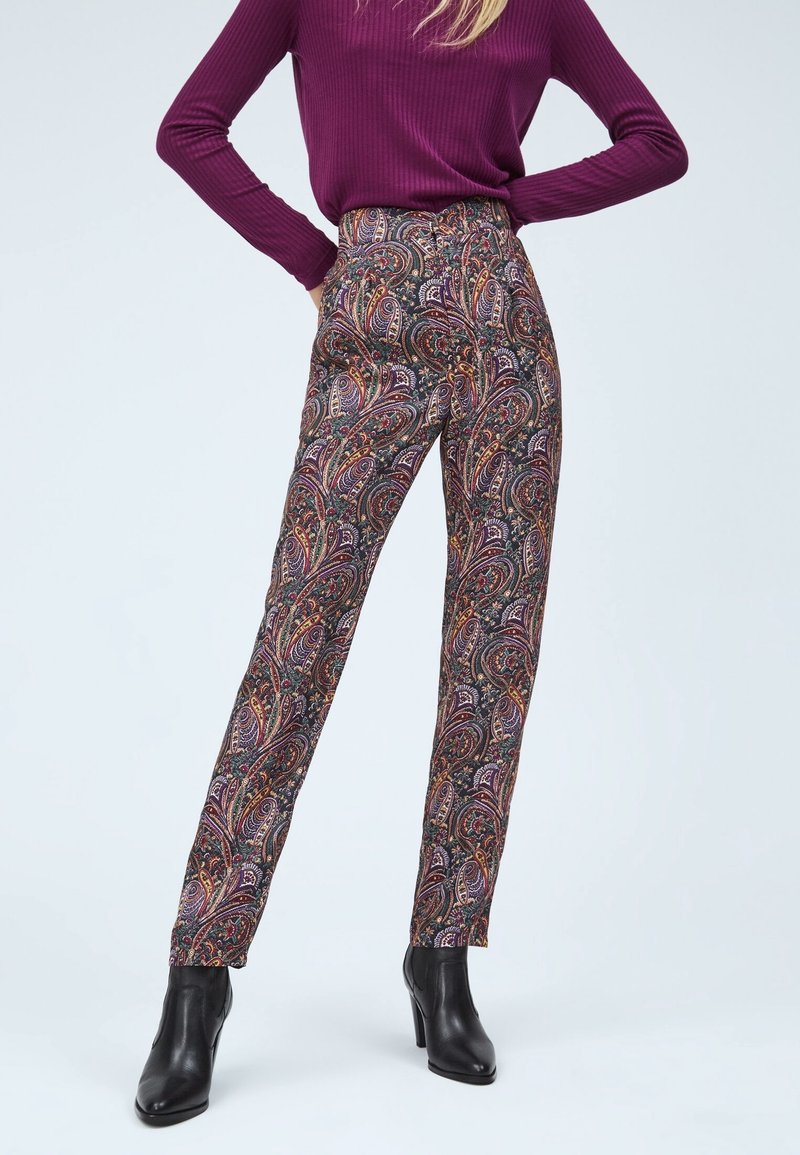 Femme portant un pantalon taille haute à motif cachemire, des bottines noires à talons et un haut violet côtelé à manches longues, debout, les mains sur les hanches.