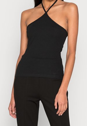 Žena nosí černý žebrovaný halter top se zkříženými ramínky a černé kalhoty, stojí před jednoduchým pozadím.