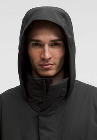 Ung mand med lys hud og kort hår iført en sort hættetrøje, der kigger direkte ind i kameraet mod en ensfarvet baggrund.