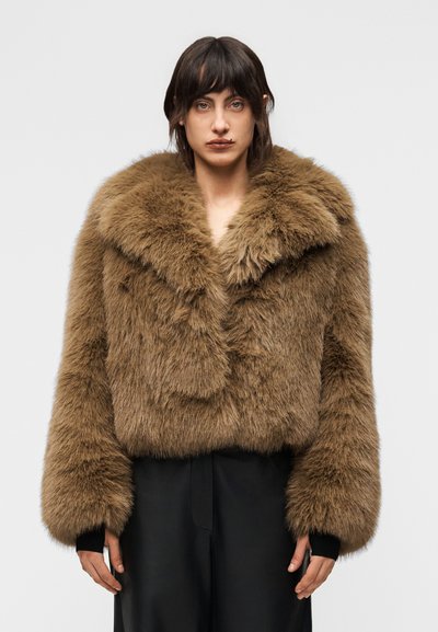 Personne portant une veste ample en fausse fourrure marron avec un grand col et des manches longues, associée à un pantalon noir, sur un fond clair uni.