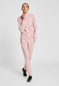 Roze trainingspak van zacht materiaal. Jas met hoge kraag, rits en witte branding; broek met zijstrepen en zakken. Zwarte sportschoenen.