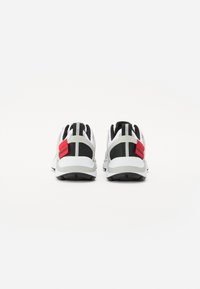 Scarpe sportive con tomaia bianca, dettagli neri, linguetta rossa sul tallone e suola texturizzata. Il dettaglio a strisce sul retro aggiunge un elemento di design.