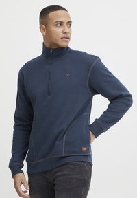 Temno modra zip-up sweatshirt iz mehkega materiala, z kontrastnim rjavim šivom, visokim ovratnikom in majhnim logotipom na prsih.