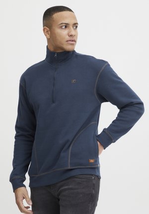 Navyblauwe ritsvest van zachte stof, met contrasterende bruin stiksel, hoge kraag en een klein logo op de borst.
