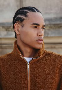 Braune Fleece-Zipjacke mit hohem Kragen, aus strukturiertem Stoff und mit einem kontrastierenden Reißverschluss. Über einem weißen Hemd getragen.