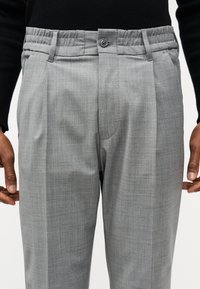 Homme portant un pantalon gris sur mesure avec fermeture à boutons et passants pour ceinture, associé à un haut noir à manches longues, les mains détendues le long du corps.