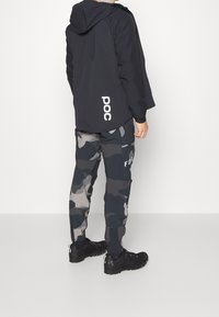 Giacca nera con cappuccio e logo bianco "POC", abbinata a pantaloni mimetici scuri con "FOX" di lato e calzature nere.