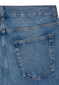 Jean en denim bleu clair avec une poche arrière rectangulaire, cousue avec un fil orange contrastant, montrant une texture lisse et un design classique.
