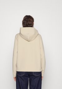 Beige mikina s kapucí a uvolněným střihem, vyrobená z měkkého materiálu. Má jednoduchý design bez viditelného kování nebo vzorů.