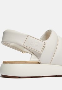 Sandal in pelle bianca con una larga fascia, soletta imbottita e suola in gomma caratterizzata da un accento marrone. Include un dettaglio con logo.