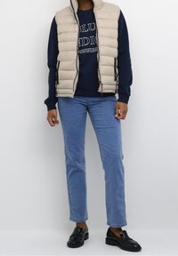 Beige gewatteerd vest over een marineblauwe sweatshirt met tekst, gecombineerd met lichtblauwe rechte spijkerbroek en zwarte loafers. Overal een gladde textuur.