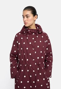 Bordowy płaszcz przeciwdeszczowy w duże białe kropki, z kapturem ze sznurkiem i elastycznym pasem. Posiada dwie boczne kieszenie oraz zapięcie na napy.