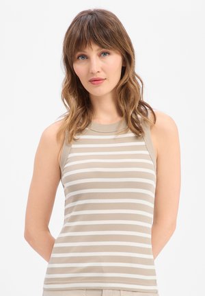 Vrouw met schouderlang golvend haar, gekleed in een mouwloos beige-wit gestreept topje, staand tegen een effen witte achtergrond.