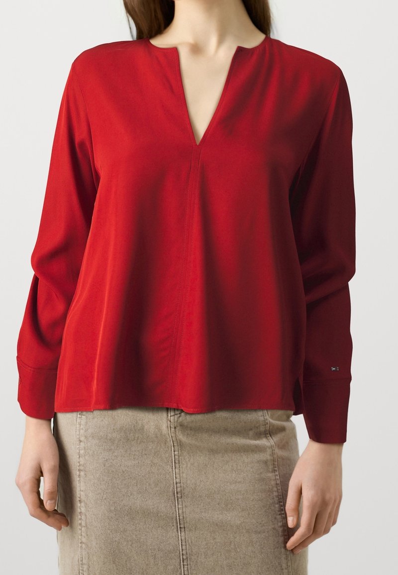 Blusa roja de manga larga con escote en V, textura suave y ligera campana en el dobladillo. Combinada con una falda beige, mostrando un estilo casual.