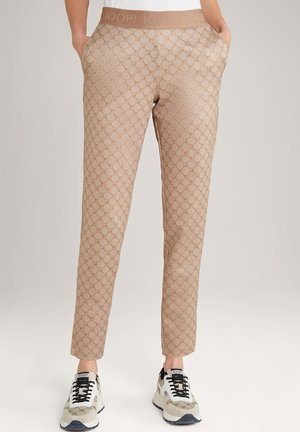 Pantalon beige fuselé avec motif circulaire répété et ceinture à logo, porté avec des baskets blanches et beiges avec lacets noirs et logo.