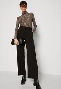 Schwarze hochtaillierte Hose kombiniert mit einem braunen und schwarzen gestreiften Langarmoberteil mit einem Stehkragen. Die Accessoires umfassen eine schwarze Clutch.