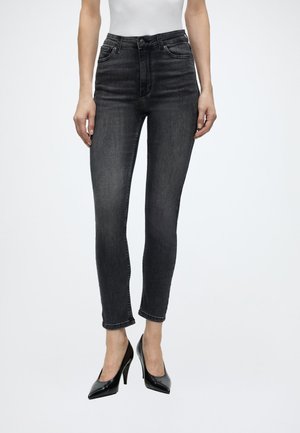 Femme portant un jean slim gris foncé et des chaussures à talons noirs, debout devant un fond blanc uni.