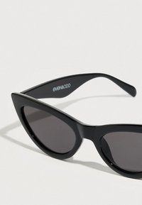 Lunettes de soleil noires à monture œil-de-chat avec des verres foncés, fabriquées en plastique lisse. Elles présentent une forme triangulaire prononcée et un logo discret sur la branche.