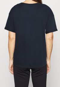 Záda osoby jsou vidět v volně padnoucím, tmavě modrém tričku, které je kombinováno s černými kalhotami, čímž se zdůrazňuje ležérní styl.