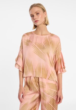 Vrouw draagt een losse roze blouse en broek met een beige palmbladerenpatroon en gerimpelde, ellebooglange mouwen, staand tegen een witte achtergrond.