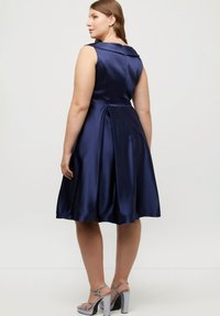 Marinblå satin klänning med figursydd byst, bred krage och full kjol. Har veck och en sidodragkedja. Matchas med silverfärgade klacksandaler.
