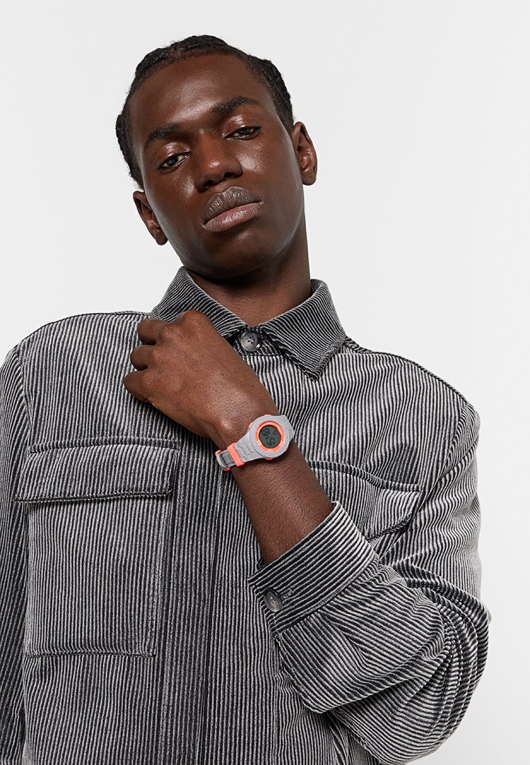 Camisa de rayas grises con cuello abotonado, que muestra un vibrante reloj digital naranja y gris en la muñeca. Enfocándose en las texturas y colores.