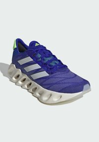 Scarpa sportiva viola con tomaia in rete, tre strisce argento e una suola intermedia bianca, testurizzata, con accenti arrotondati. Dettaglio del tallone verde.