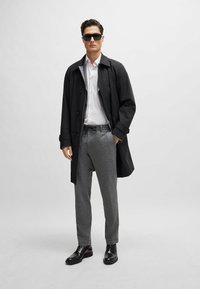 Musta trench coat valkoisen paidan päällä, harmaa kuvioitu housut ja mustat kengät. Asusteina tummat aurinkolasit. Sulavat tekstuurit, mukautettu leikkaus.