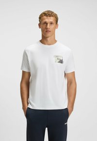 Hvid kortærmet T-shirt i blød bomuld. Har et stofmærke på brystet med ordet "BOSS" og abstrakte designelementer.