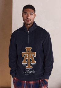 Marineblauer Fleece-Pullover mit Viertel-Reißverschluss, der ein strukturiertes, beiges Logo mit den Schriftzügen "A.T." und "Austin" zeigt. Weicher Stoff und lockere Passform mit langen Ärmeln.