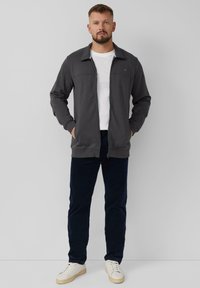 Giacca grigia zip-up con colletto, t-shirt bianca sotto, abbinata a pantaloni blu scuro e sneakers bianche. Design semplice e casual.