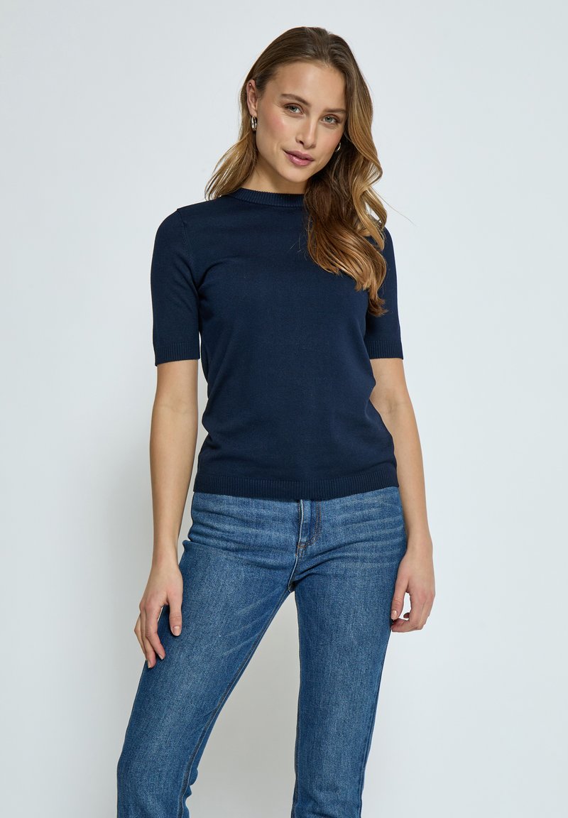 Marinblå stickad kortärmad topp med rund halsringning och ribbade kanter, kombinerad med medelblå jeans, vilket skapar en figursydd silhuett.