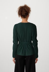 Persona con capelli ricci che indossa un top a maniche lunghe a coste verde scuro con una cintura in vita, girata di spalle, abbinato a pantaloni neri.