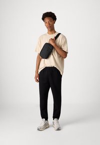 Bandolera negra hecha de material duradero, llevada sobre una camiseta beige y pantalones de chándal negros. Presenta una textura suave y un cierre de cremallera seguro.
