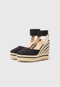 Espadrilles à talon compensé en toile noire avec une semelle en jute tressé, des lacets en tissu noir entrecroisés à la cheville et des détails en jute rayé le long du talon.