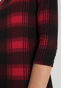 Černá a červená kostkovaná košile s měkkou texturou, zobrazující zaoblený design rukávů a kontrastní horizontální a vertikální vzory.