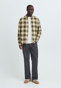 Rutete flanellskjorte i oliven og beige, knappet, brukt over en hvit T-skjorte. Kombinert med mørkegrå jeans med rett ben og beige sneakers.