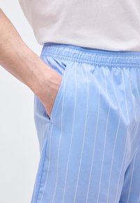 Χέρι στην τσέπη ανοιχτόμπλε παντελονιού με λευκές κατακόρυφες ρίγες και ελαστική μέση, συνδυασμένο με λευκό πουκάμισο.