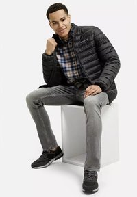 Czarna kurtka puchowa o strukturalnym wzornictwie, w połączeniu z koszulą w kratę, szarymi dżinsami i czarnymi butami sportowymi, siedząca na białym sześcianie.