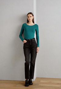 Langsleeve teal shirt gecombineerd met hoge zwarte jeans. Model draagt zwarte schoenen, staat tegen een lichtgrijze muur op een houten vloer.
