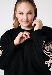 Zwarte trui met opgeblazen mouwen en beige bloemrijke borduursel op de armen. Het model draagt ringen en past de mouwzoom aan.