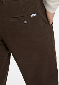 Bruna kordbyxor med vertikal ribbing, som har en liten varumärkesetikett och en knappstängning på frampocket.